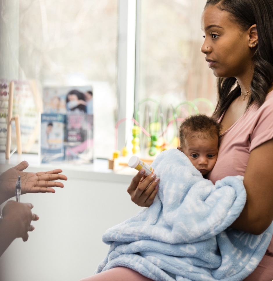 The serious new young adult mother holds her baby to her chest as she listens to the unrecognizable doctor describe possible side effects of a new medication.