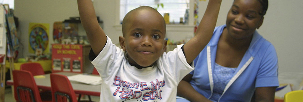 child in head start program with a child care worker in the background smiling and playing in a child care center